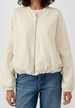 Vrouw draagt een licht beige jas met ritssluiting en elastische manchetten en zoom, gecombineerd met blauwe spijkerbroek tegen een egale achtergrond.