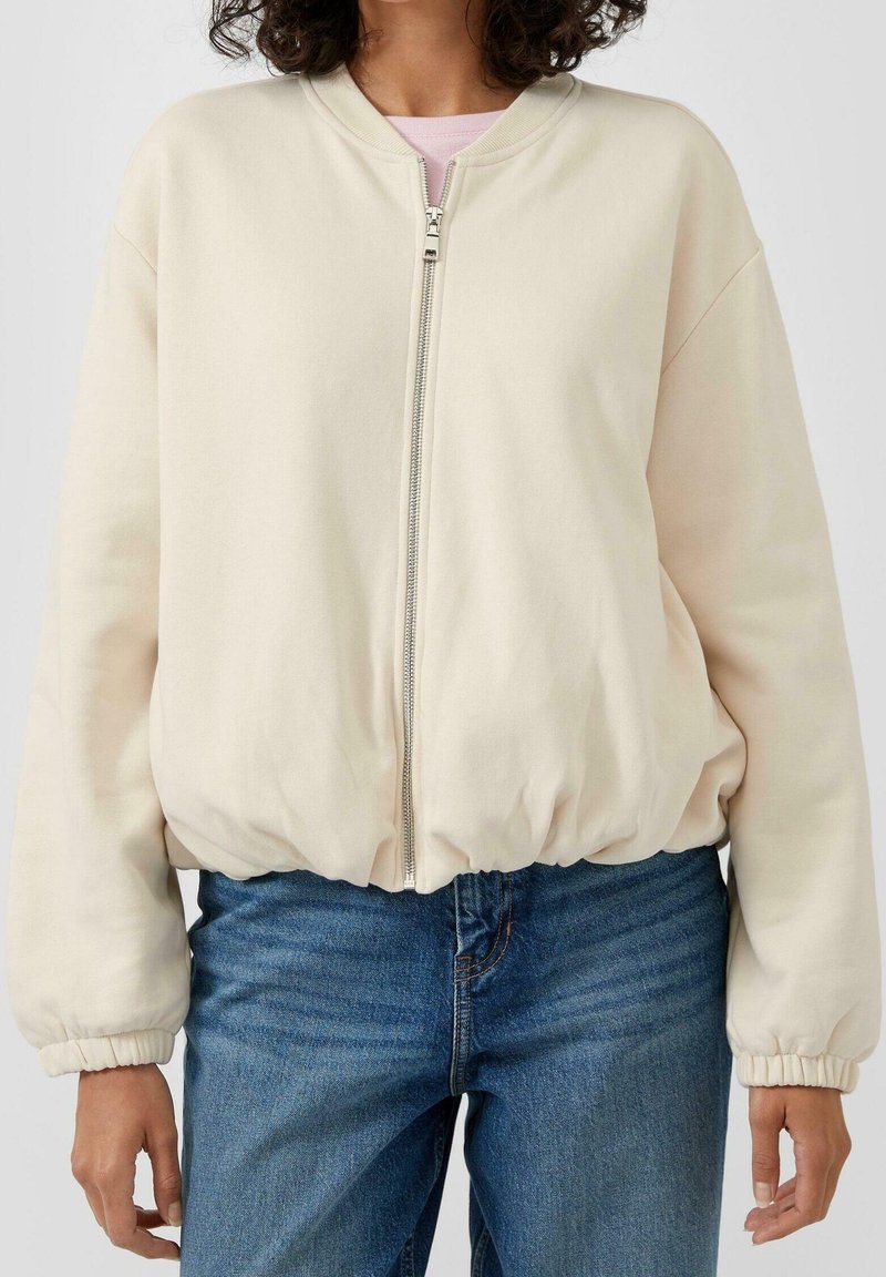 Femme portant une veste zippée beige clair avec poignets et ourlet élastiques, associée à un jean bleu sur un fond uni.