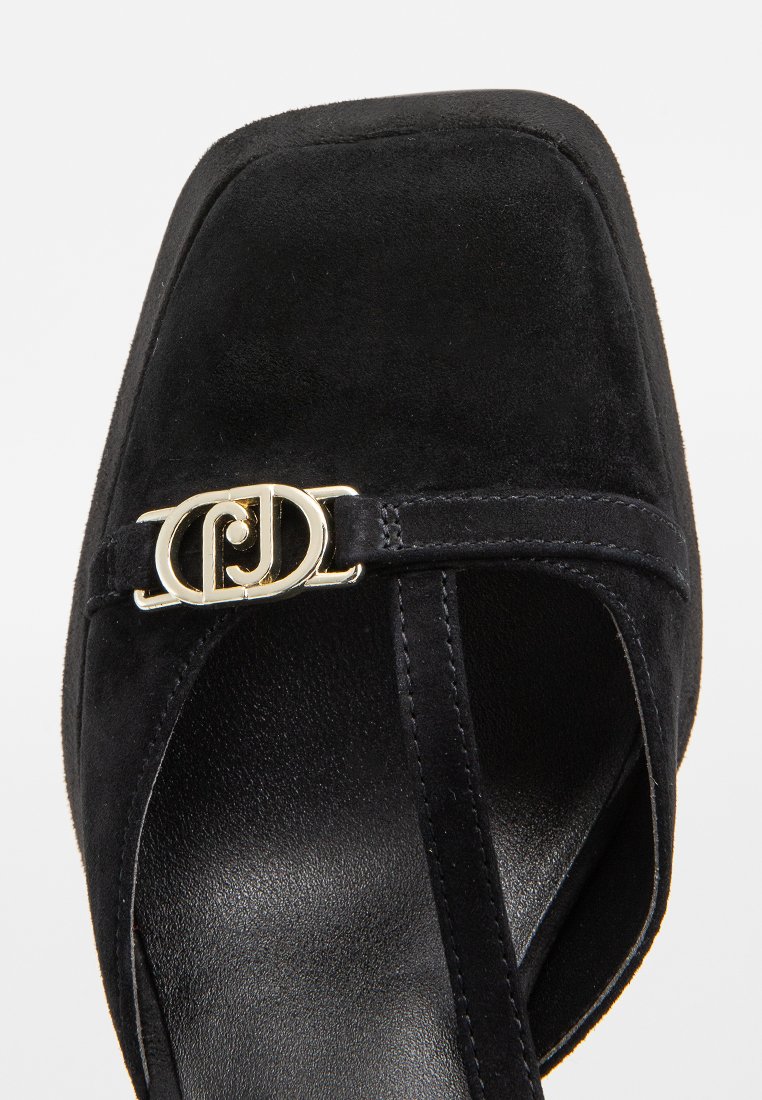 Zapato de tacón alto de ante negro con punta cuadrada, tira estrecha y herrajes con logo dorado en la parte delantera. Acabado elegante y textura suave.