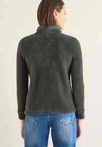 Tmavě zelený fleecový svetr s měkkou texturou a složeným límcem, doplněný světlými modrými džínami. Pohled zezadu zdůrazňující délku rukávů.