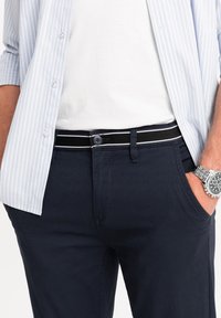 Pantaloni blu navy con una cintura a righe bianche e nere, abbinati a una t-shirt bianca e una camicia a righe azzurre, con un orologio d'argento.