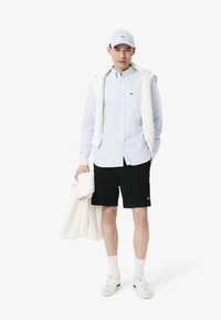 Lichtblauw overhemd, zwarte shorts, witte sweater over de schouders, witte sneakers en een bijpassende pet. Lacoste-logo op het overhemd en de shorts.