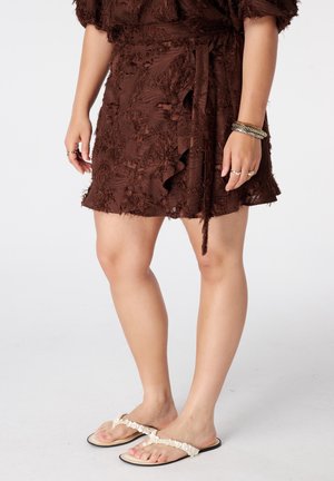 Vrouw draagt een bruine, getextureerde wikkelrok met riem, witte platte sandalen met schelpenversiering en armbanden om haar rechterpols.