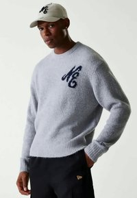 Γκρι πλεκτό πουλόβερ με στρογγυλή λαιμόκοψη και μπλε σκούρο λογότυπο στο στήθος. Φορεμένο με μπεζ καπέλο και μαύρο παντελόνι, παρουσιάζοντας έναν καθημερινό, χαλαρό στιλ.