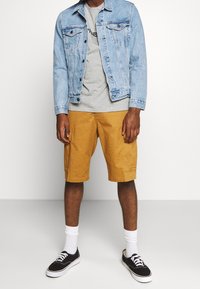 Lichtblauwe denimjasje over een grijze shirt, gecombineerd met gele shorts en zwarte sneakers. Zichtbare witte sokken. Eenvoudig ontwerp, casual uitstraling.