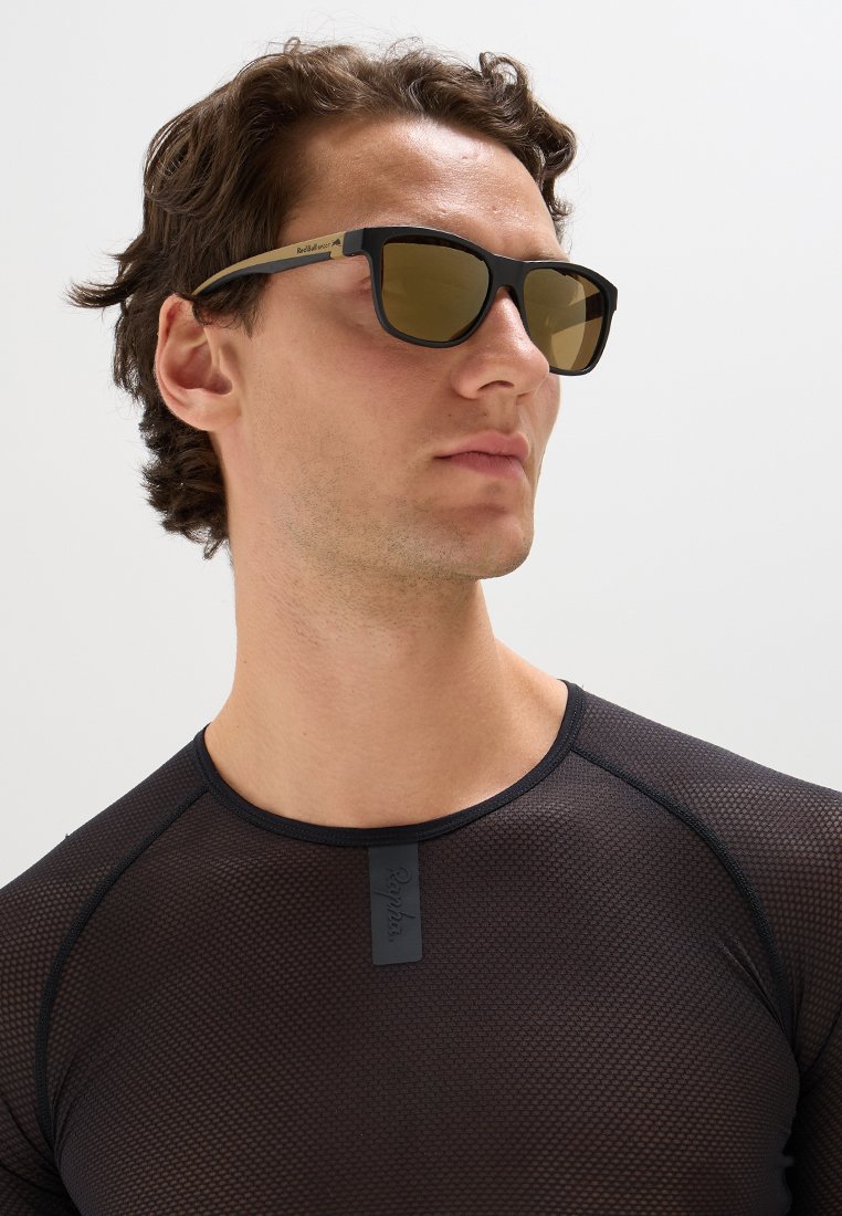 Czarne okulary przeciwsłoneczne z przyciemnionymi brązowymi szkłami, z matową czarną oprawką. Modelka ma na sobie czarną, siatkową bluzkę z długim rękawem.