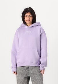 Lavendel hettegenser med frontlomme, ribbestrikkede mansjetter og nederkant, med "STUDIO EDITION"-tekst trykt i hvitt på brystet.