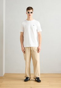 Hvid kortærmet t-shirt med lille sort logo på brystet, parret med beige brede bukser og sorte sneakers.