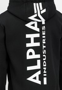 Capuz preto feito de algodão, apresentando um grande logótipo branco "ALPHA INDUSTRIES" verticalmente nas costas, com um acabamento de tecido texturizado.