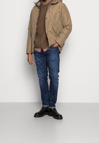 Giacca a piumino marrone chiaro con cappuccio, maglione marrone lavorato a maglia, orlo di camicia a quadri, jeans blu e stivaletti neri alla caviglia, creando un look casual a strati.