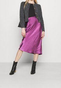 Černý kardigan z umělé kožešiny se zapínáním na knoflíky, doplněný lesklou růžově fuchsiovou saténovou sukní po kolena a černými kotníkovými botami na blokovém podpatku.