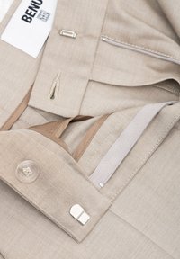 Beige stoffen broek met een gladde textuur. Kenmerken zijn een knoop, een ritssluitingzak en geweven accenten in een lichtere tint. Label zichtbaar.