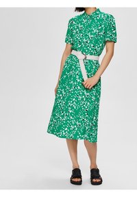 Abito floreale verde con maniche corte, collo a polo, vita con cintura e lunghezza midi. Il design presenta motivi di foglie bianche.