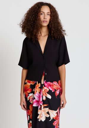 Donna con i capelli ricci che indossa una camicia nera a maniche corte con bottoni e una gonna nera con grandi motivi floreali rosa e arancioni.