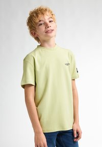 Lichtgroene T-shirt met korte mouwen en een ronde hals, voorzien van een klein zwart logo op de borst, gecombineerd met donkerblauwe jeans.