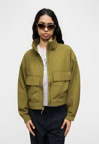 Γυναίκα που φοράει χακί ζακέτα με φερμουάρ και μεγάλες μπροστινές τσέπες, λευκό μπλουζάκι με τύπωμα, μαύρο παντελόνι και γυαλιά ηλίου cat-eye μπροστά σε απλό φόντο.