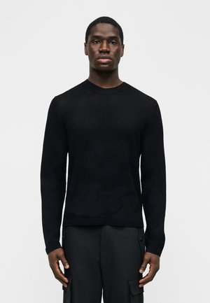 Mlad moški stoji obrnjen naprej, oblečen v črno dolg rokav pulover in črne cargo hlače, na enostavni svetli podlagi.