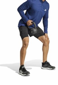 Černé sportovní šortky a modré tričko s dlouhým rukávem a půlzipem. Osoba drží černou kettlebell a má na sobě černé tréninkové boty.