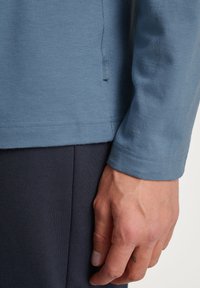 Lichtblauw langelijfshirt van zachte stof, met een subtiele textuur, een afgeronde zoom en een klein merketiket aan de zijkant.