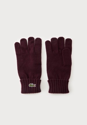 Par de guantes de punto de color púrpura oscuro con puños de canalé y logo verde de cocodrilo en el puño del guante izquierdo, colocados planos sobre fondo blanco.