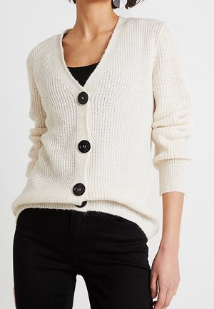 Femme portant un cardigan en tricot blanc avec trois gros boutons noirs sur un haut noir et un pantalon noir.
