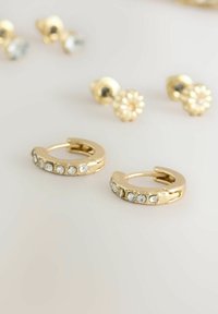 Orecchini a cerchio in oro con una fila di strass trasparenti lungo il bordo anteriore. In secondo piano, ci sono orecchini a stud con design floreali.