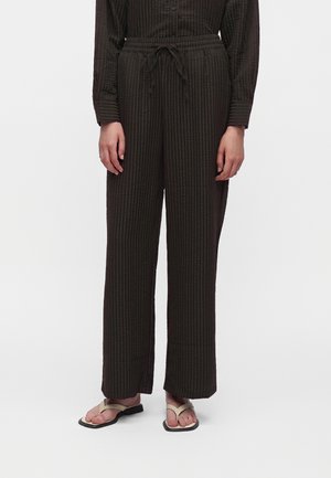Personne portant un pantalon large texturé marron foncé avec une taille à cordon et des sandales beige contre un fond uni.