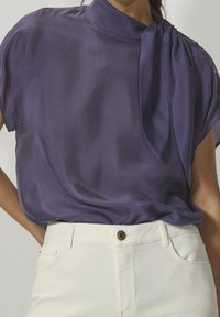 Donna che indossa una blusa di seta viola con un dettagli al collo a cravatta e pantaloni bianchi a vita alta, in piedi con una mano sulla coscia.