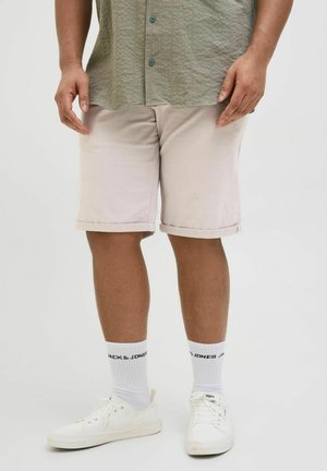 Pantaloni scurți de culoare bej deschis cu tivuri rulate, combinați cu adidași albi și șosete care afișează textul "JACK & JONES" în negru.