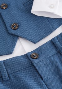 Chaleco azul tejido con solapa en pico, adornado con botones marrones. Se asoma el puño de una camisa blanca debajo. Corte entallado con texturas suaves.