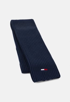 Écharpe en laine tricotée bleu marine, pliée, avec une petite étiquette rectangulaire en tissu rouge, blanc et bleu près d'un bord.