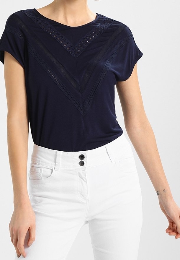 Kvinna iklädd en marinblå topp med spetsdetaljer som formar en V-form, ihop med högmidjade vita byxor som fästs med två knappar.