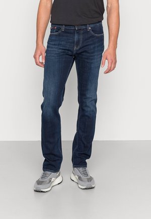 Man wearing dark blue slim-fit jeans and gray and white sneakers standing against a white wall and gray floor.