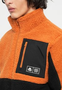 Sudadera de polar naranja con un bolsillo negro con cremallera, que presenta una superficie texturizada y acentos en negro. Parche del logo en el pecho.