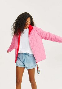 Prešívaná bunda v ružovej a červenej farbe, s vysokým golierom, plným zipom a dlhými rukávmi. Nosená cez bielu košieľu a svetlomodré džínsové šortky.