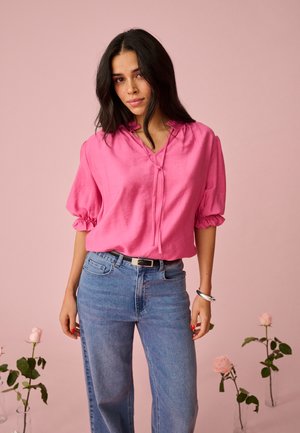 Frau mit langen dunklen Haaren, die eine leuchtend pinke Bluse mit Puffärmeln und hellblaue Jeans trägt, steht in der Nähe von Glasvasen mit rosa Rosen.