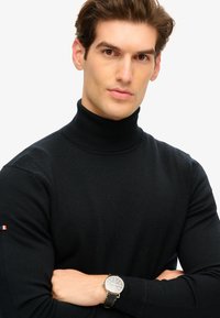 Maglione a collo alto nero in un design a costine, con un sottile dettaglio del logo sulla manica sinistra e un orologio argentato con cinturino nero.