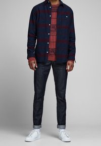 Hemd mit Knopfleiste in Marineblau und Bordeaux gestreift, über einem rot gemusterten Pullover getragen. Kombiniert mit dunklen Jeans und weißen Turnschuhen.