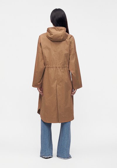 Femme debout de dos, portant un long manteau marron à capuche et un jean bleu à jambes larges avec des ourlets effilochés, sur un fond uni.