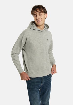 Ung mand smilende iført en lysegrå hættetrøje med lille logo på brystet og blå jeans mod en ensfarvet baggrund.