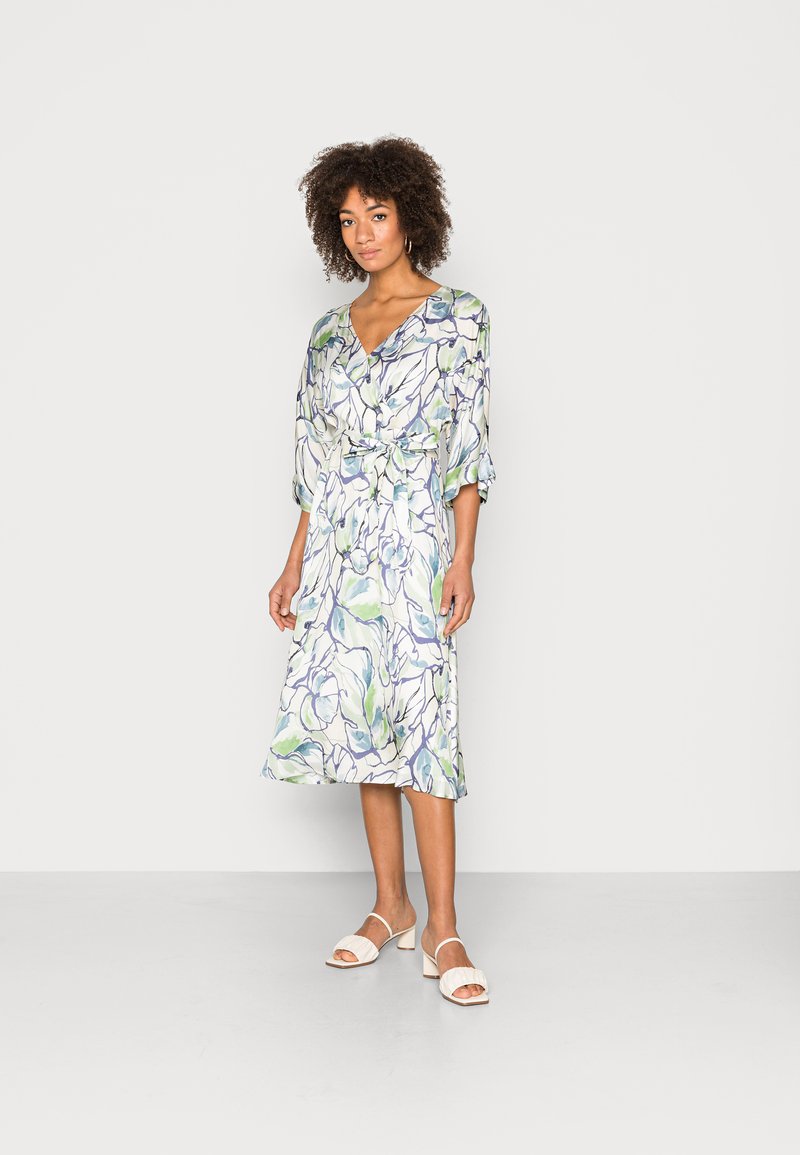 Vestido com padrão floral, decote em V, mangas de três quartos e um laço na cintura. As cores incluem verde, azul e branco. Combinado com sandálias leves.