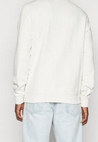 Persona vista da dietro che indossa un maglione bianco a maniche lunghe semplice e jeans di denim blu chiaro con tasche posteriori visibili.