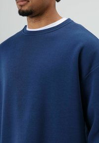 Marineblauwe sweater gemaakt van zachte stof met een ronde halslijn en geribbelde details. Heeft afhangende schouders en een ontspannen pasvorm.