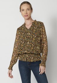 Blusa nera floreale con fiori gialli e arancioni, scollatura a V, maniche lunghe con polsini arricciati e orlo elastico. Tessuto leggero e trasparente.