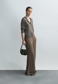 Cardigan marron avec des boutons, associé à un pantalon marron à jambes larges. Le modèle tient un sac à main vert foncé, sur un fond neutre.
