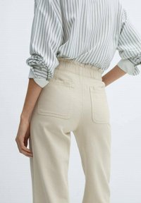 Pantaloni beige a vita alta con elastico in vita, due tasche posteriori e una vestibilità comoda, abbinati a una camicia a maniche lunghe a righe.