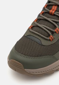 Zelená turistická obuv se strukturovaným síťovaným svrškem, gumovou podrážkou a oranžovými akcenty na šněrování. Obsahuje polstrovaný límec a zesílenou špičku.