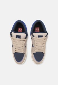 Sneakers beige e blu navy con una tomaia in tessuto testurizzato, dettagli in contrasto e lacci piatti. Soletti brandizzati rossi visibili all'interno.