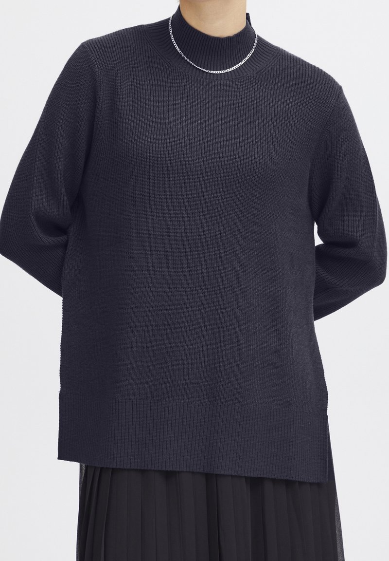 Pull marine foncé avec une texture côtelée, col montant et coupe ample. Il présente un ourlet droit et des manches arrivant aux coudes.