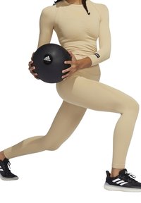 Maglietta sportiva beige a maniche lunghe con tessuto texturizzato, abbinata a leggings coordinati. Tiene in mano una palla medica nera con il logo Adidas.
